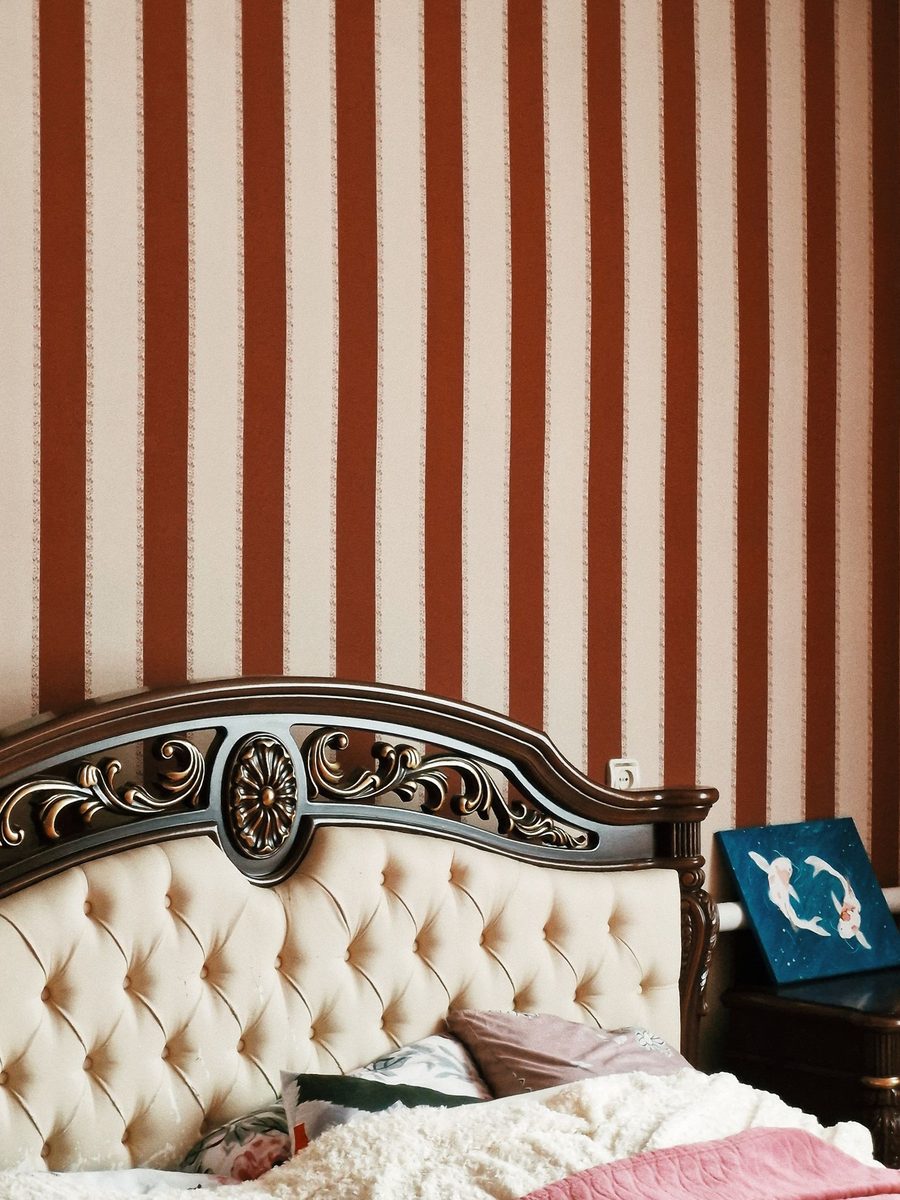 Bedroom with vertical rust-and-cream striped wallpaper above a tufted, carved headboard