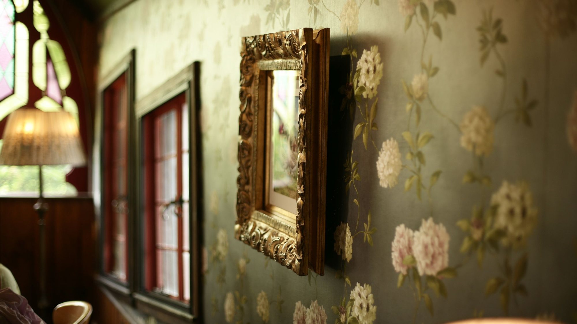 Elegant interior with soft sage floral wallpaper, an ornate gilt-framed mirror and warm natural light from a stained-glass window
