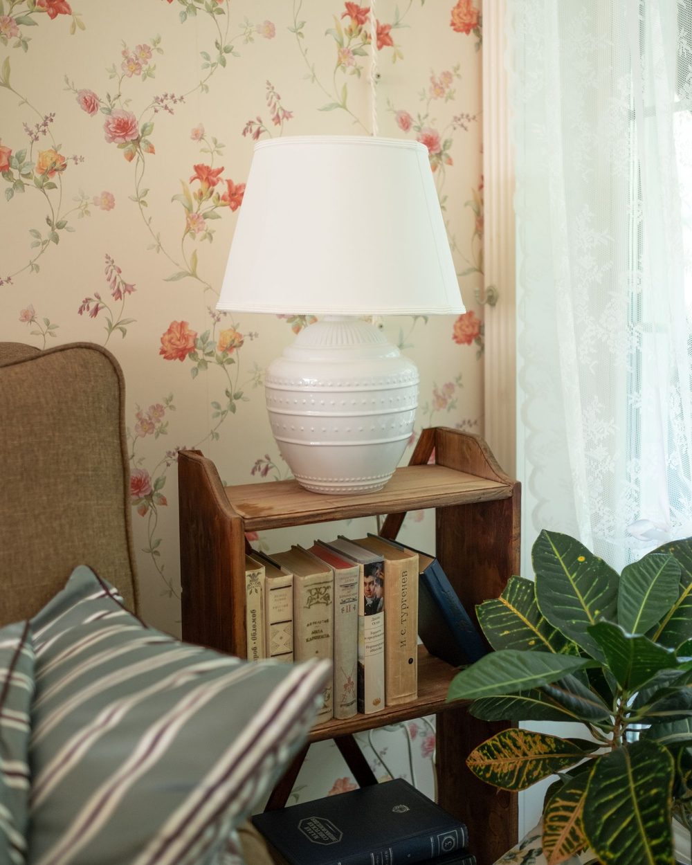 Warm interior corner with floral wallpaper, a lit table lamp on a wooden shelf and a green plant